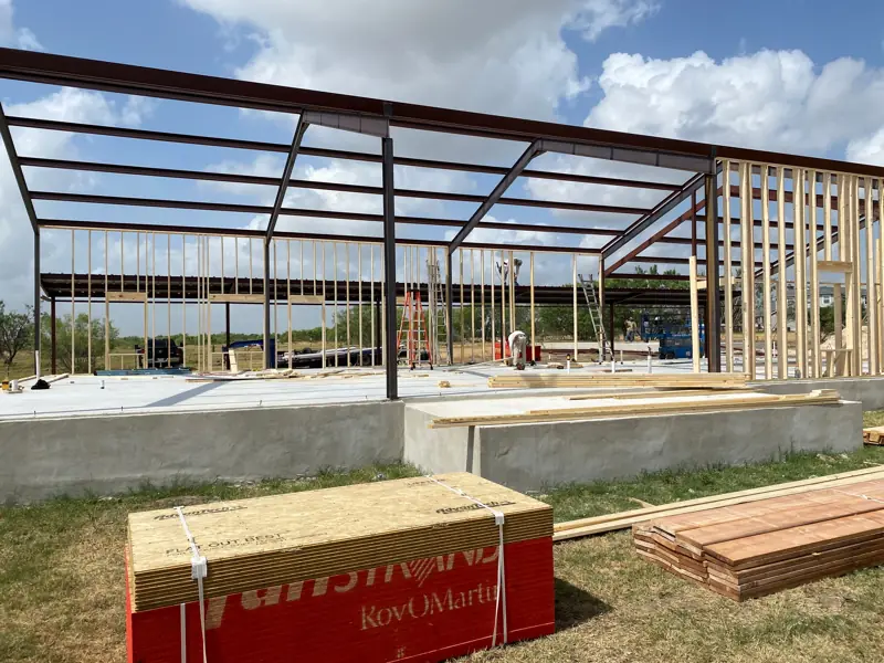 Steel frame on a raised slab with wood wall framing starting, lumber and sheathing on the ground.