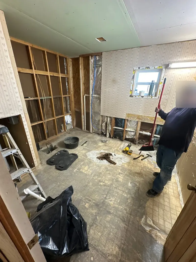 Bathroom with the walls opened to the studs and the old floor still down, the plumbing showing and a worker sweeping up.