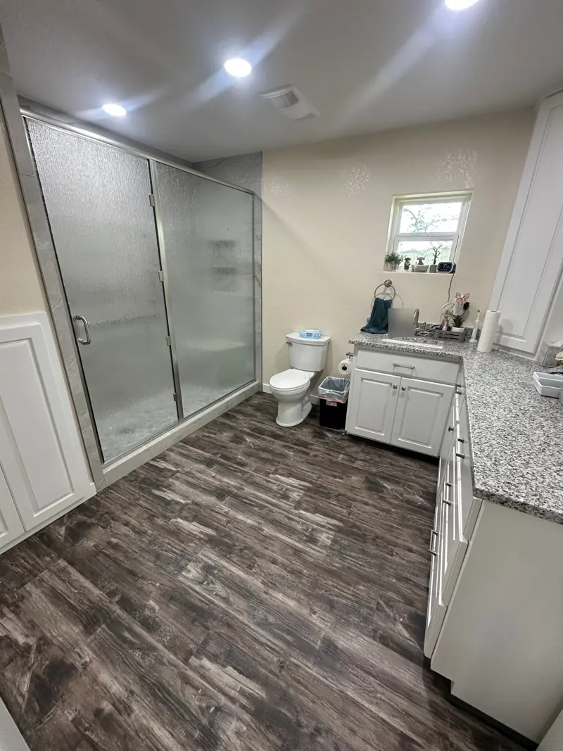 Bathroom with textured glass shower doors, a long white vanity with a speckled stone top and recessed lights.