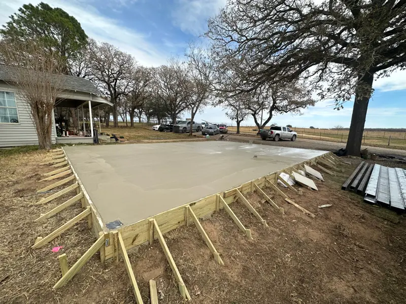 Freshly poured and finished concrete slab inside braced wood forms, beside the house on open ranch land.