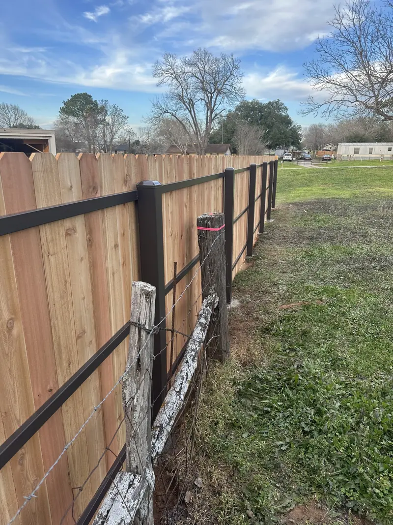 Cedar dog-ear pickets on dark metal posts and rails, running beside an old barbed wire fence.