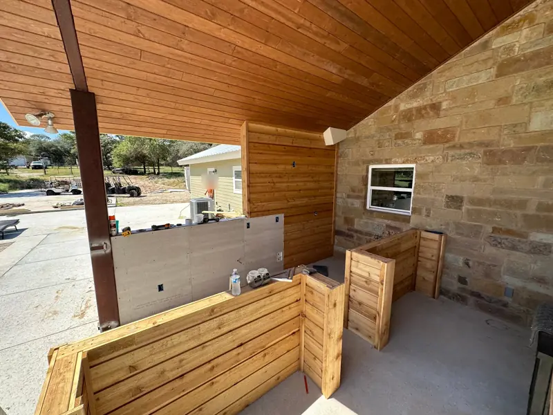 Cedar cabinet bases for an outdoor kitchen under a porch with a cedar ceiling and a stone wall.