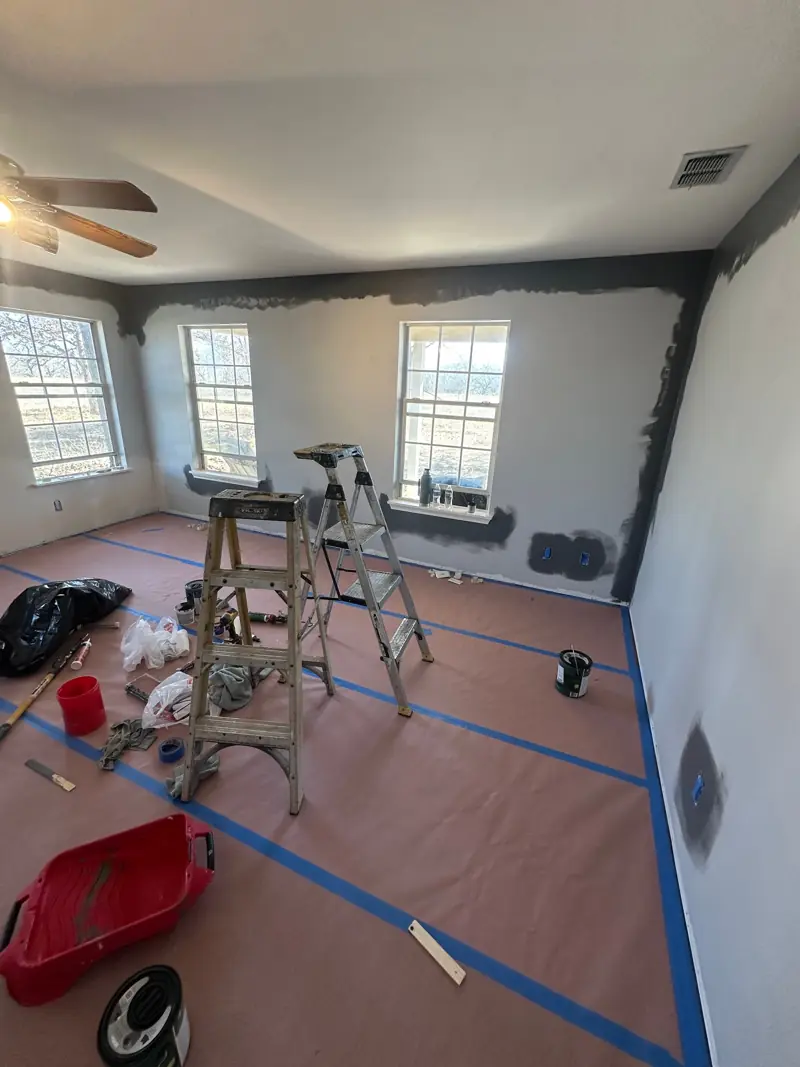 Dark gray paint cut in around the bedroom windows, corners and ceiling, with ladders and paint supplies on the covered floor.