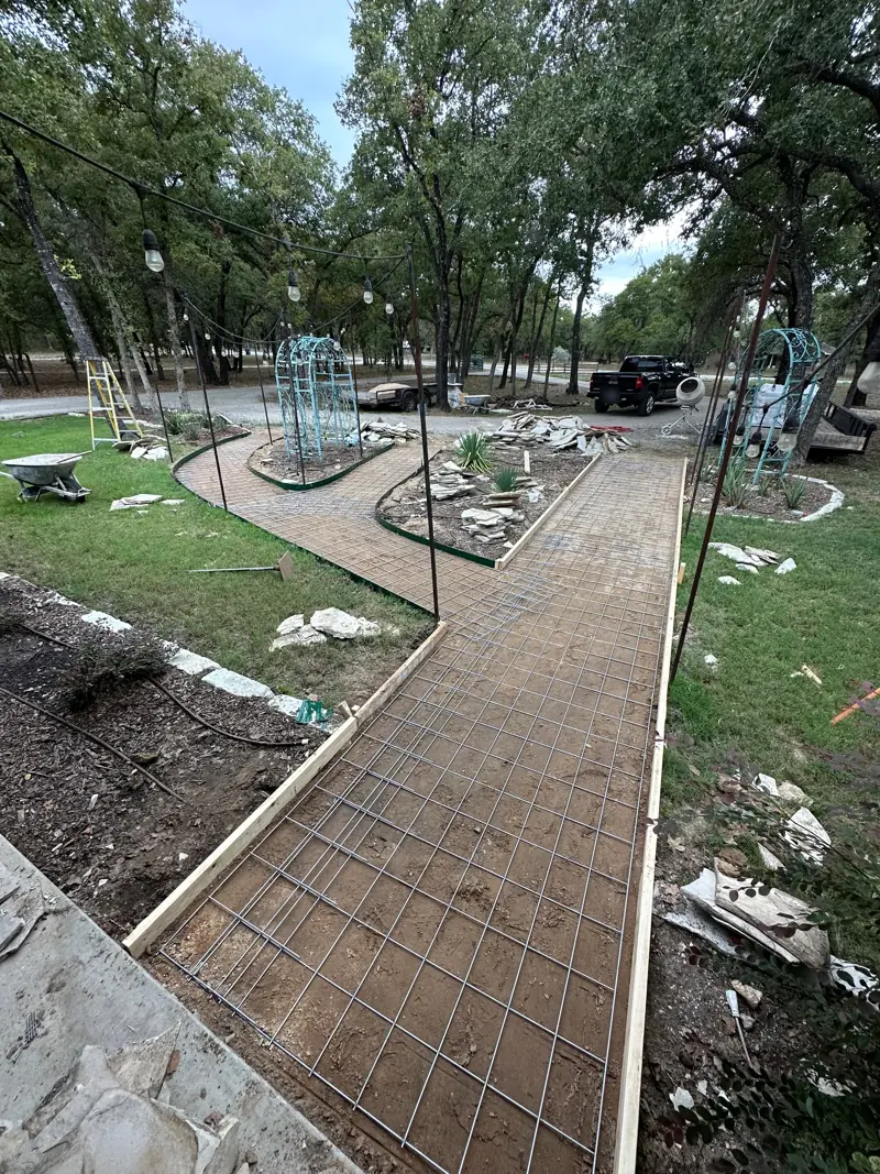 Walkway formed with wood boards and rebar mesh, leading between garden beds with metal arbors under oak trees.