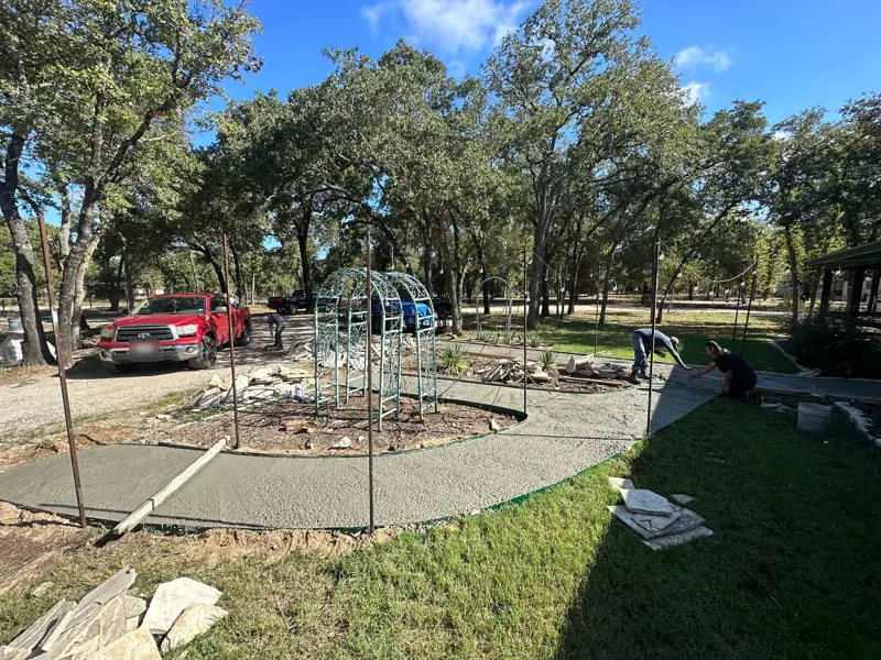 Crew finishing a curved concrete base around a garden bed with a metal arbor, flagstone stacked beside it.