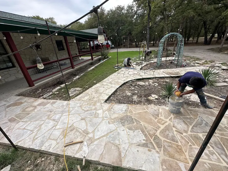 Crew setting flagstone on the concrete base around the garden beds, beside a covered porch with red posts.