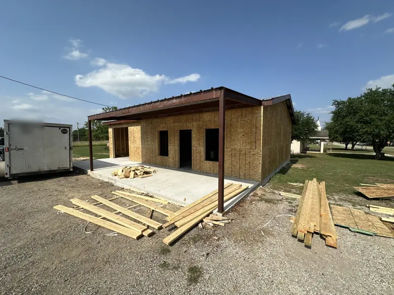 The building in OSB sheathing, with a steel lean-to porch roof over a new slab.