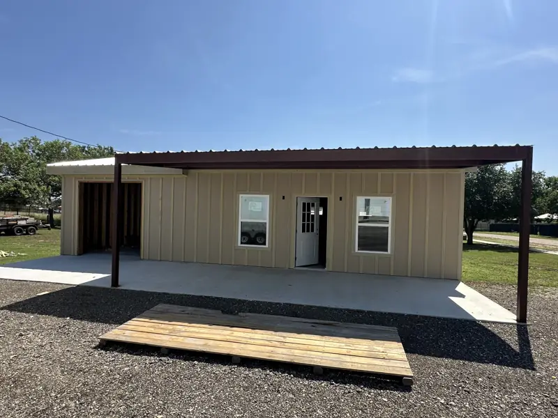 Finished building with tan siding, white windows and door, and a covered porch on steel posts.