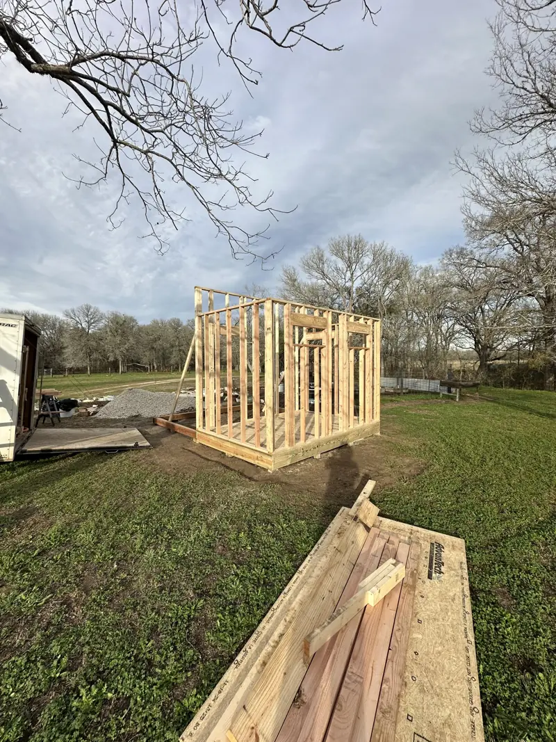 Wood stud walls framed on the shed floor deck, with lumber stacked on a subfloor sheet in the foreground.