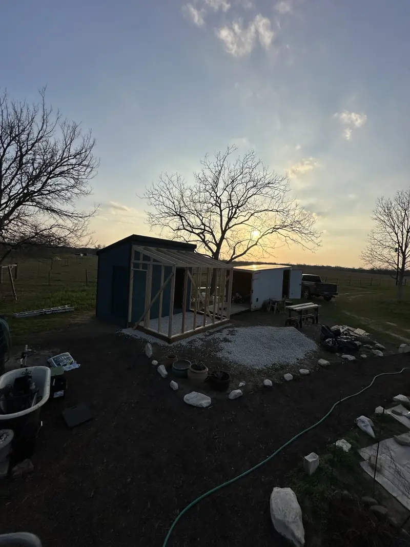 Dark blue shed at sunset with wood framing for an attached greenhouse built over a white gravel floor.