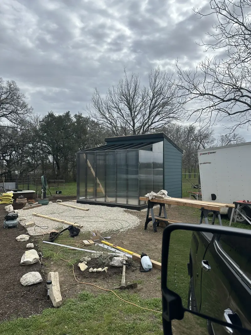 Greenhouse with clear polycarbonate panels attached to a blue shed, with tools and lumber on the ground.