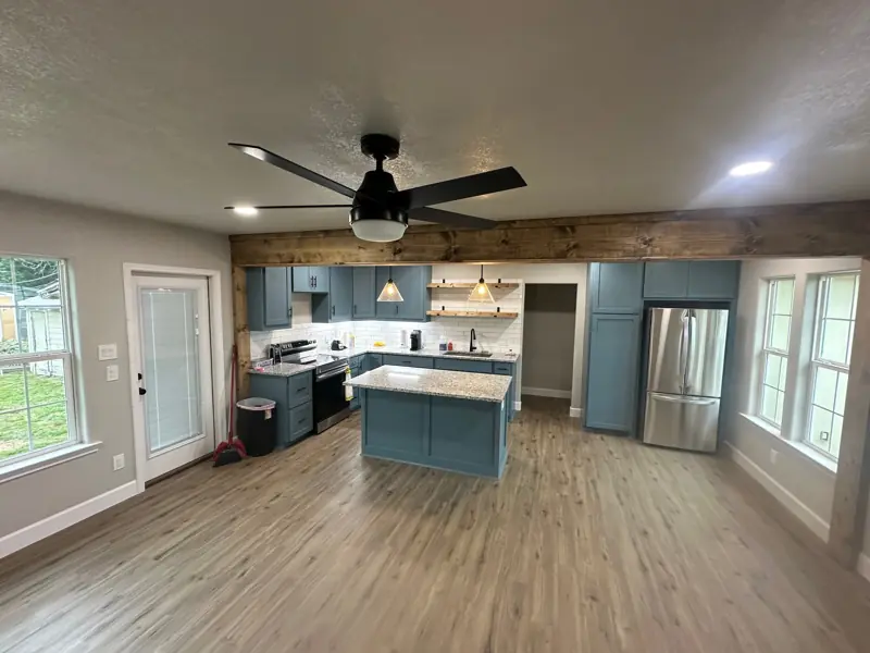 Finished open living room and kitchen with wood-look plank floors, blue cabinets, an island, a wood beam and a ceiling fan.