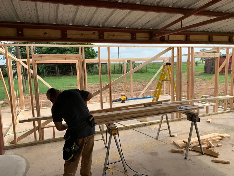 Worker cutting lumber under a metal roof, new wall framing behind.