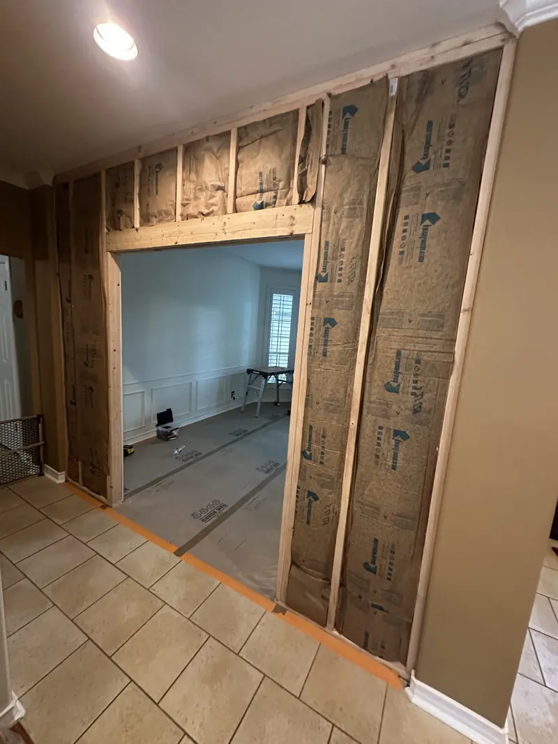 Framed wall with kraft-faced insulation and a wide door opening into a dining room with protected floors.