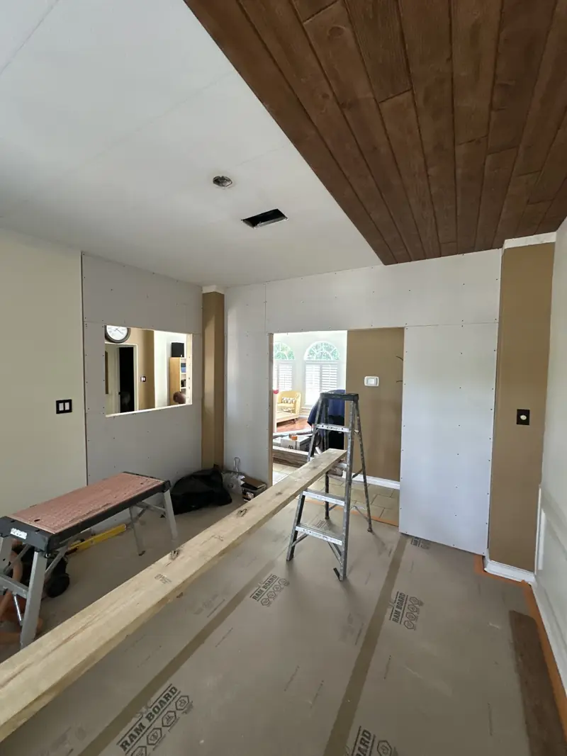 New drywall on the room's two new walls, with a window cutout, a doorway and a stained wood plank ceiling above.