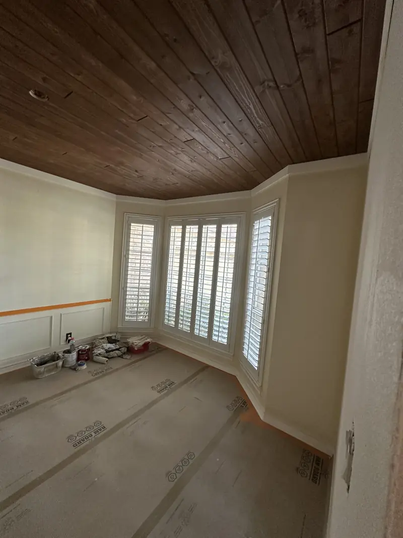Stained wood plank ceiling over a bay window with plantation shutters, with paint supplies on the protected floor.