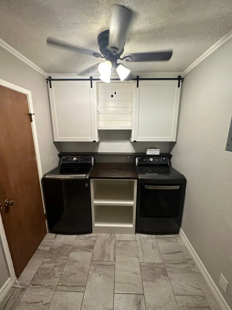 Utility room with white cabinets with sliding barn-style doors over the washer and dryer, open shelves between and a tile floor.