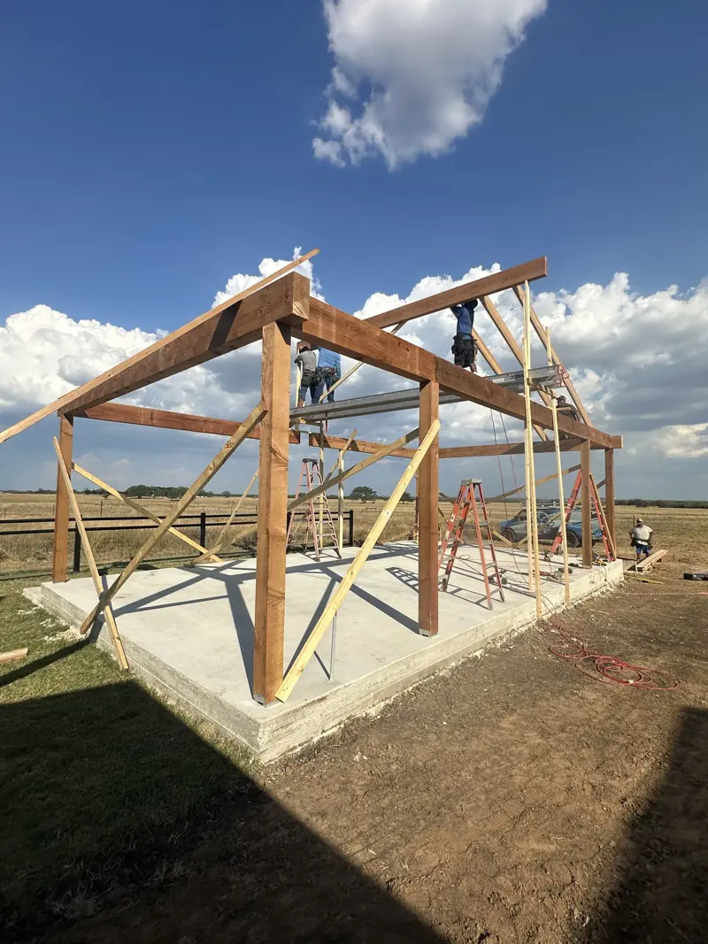 Rough-sawn timber posts and beams with temporary diagonal bracing on a concrete slab, crew working on scaffold above.