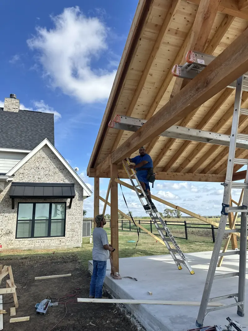 Carpenter on an extension ladder working under a new wood plank roof deck while a crew member watches from the slab.