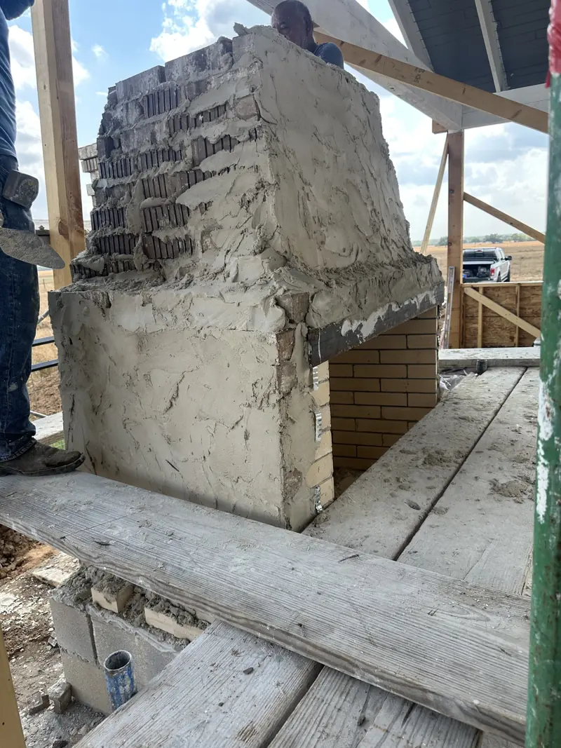 Fireplace under construction with brick firebox and stack in a rough mortar coat, scaffold planks in front.