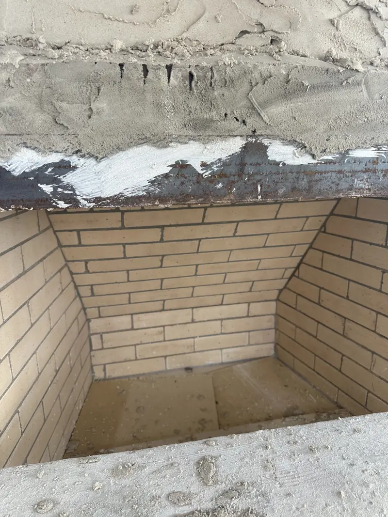 Close-up of a firebox lined with tan firebrick, with angled side walls under a steel lintel and fresh mortar above.