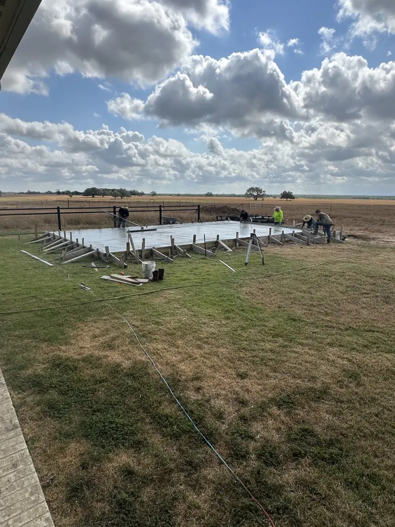 Crew finishing a large concrete slab in an open field beside a ranch fence.