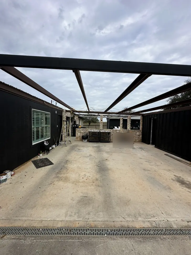 Patio roof stripped to its steel beams, open sky above the concrete slab between black-sided walls.