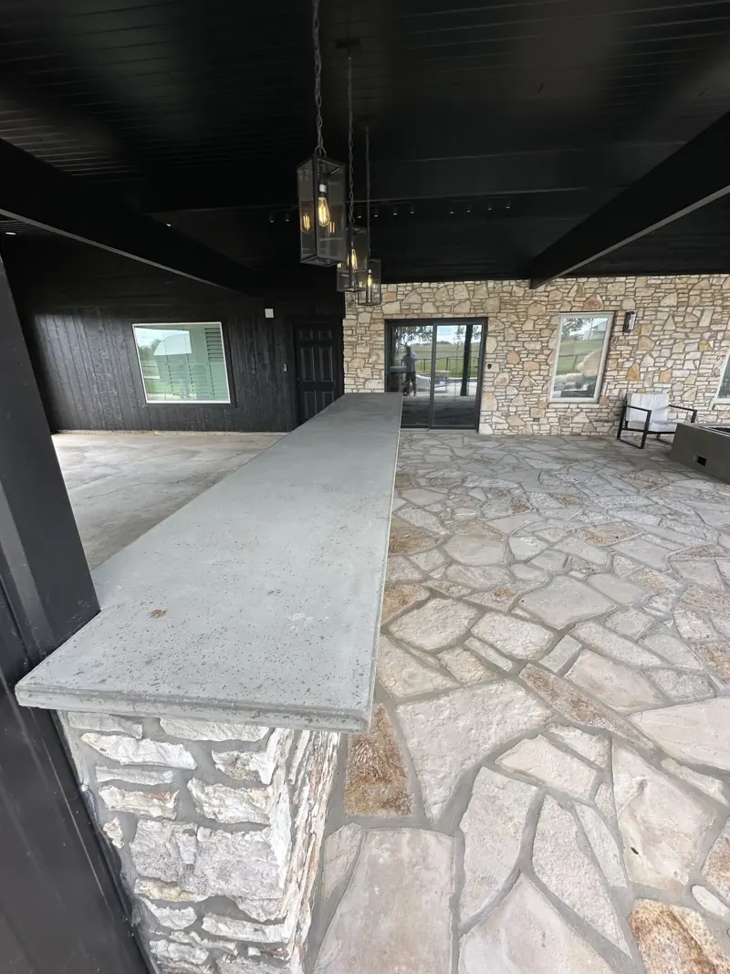 Concrete bar countertop on a stone wall under black ceiling beams, with glass lantern pendants overhead.