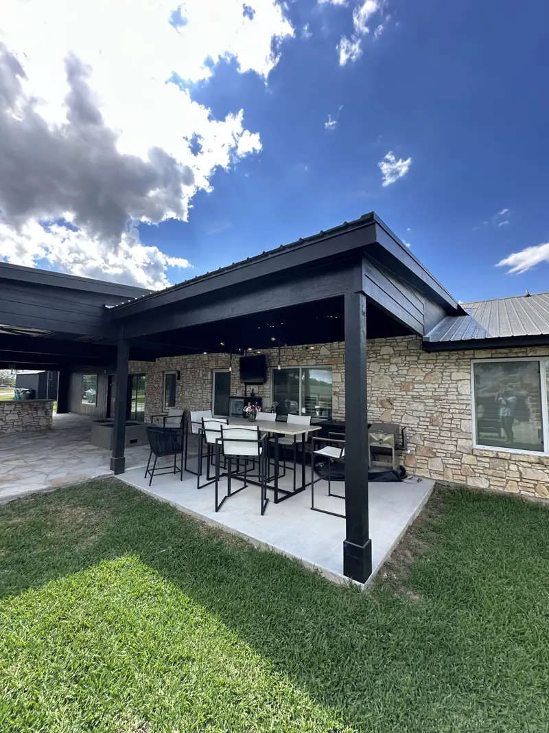 New covered dining area on a concrete slab, black posts and metal roof against the stone house, blue sky.