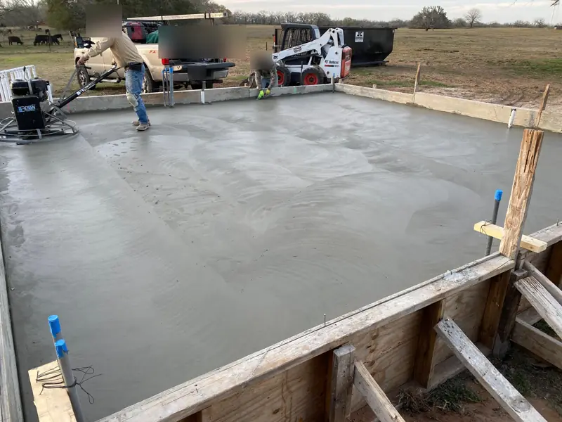 Crew running a power trowel over a fresh slab, a skid steer at the far edge.