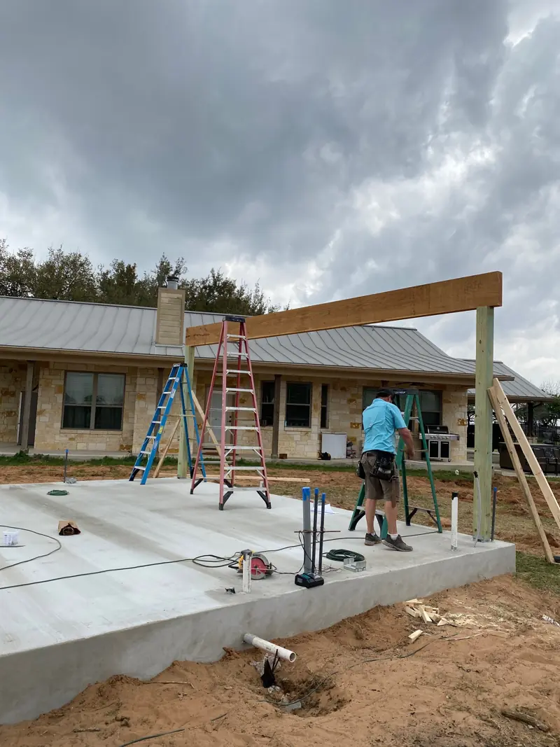 First cedar posts and a beam going up on the new slab beside a stone house.