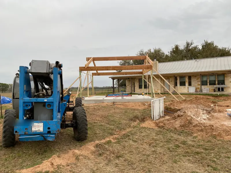 Telehandler beside the cedar frame on the slab, the stone house behind.