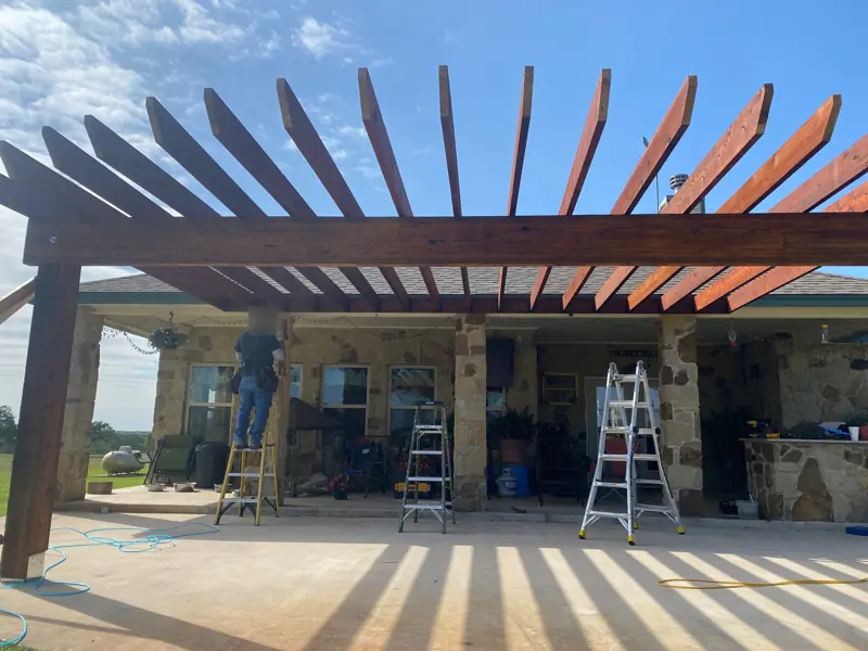Curved cedar pergola going up over the patio of a stone house, a worker on a ladder.
