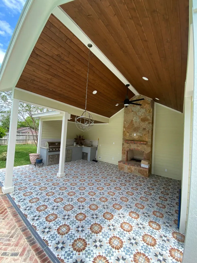 Covered patio with a stained wood ceiling, patterned tile floor and a stone fireplace.