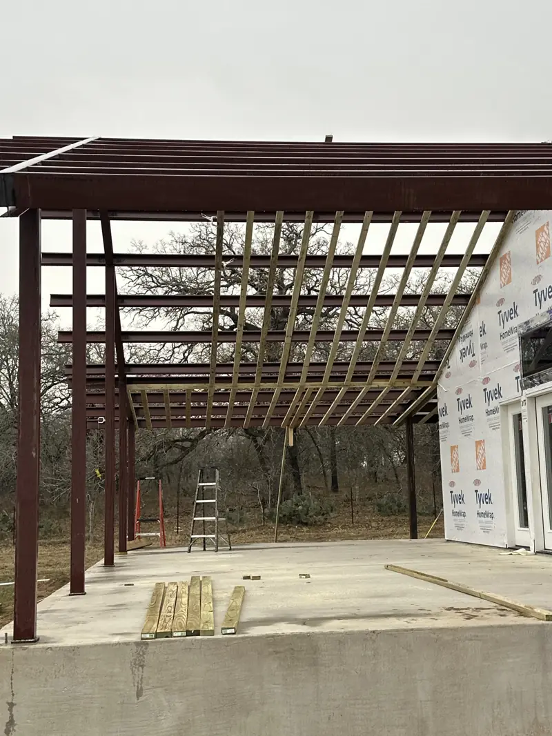 Steel posts and beams with wood purlins over a concrete slab, built against a house in house wrap.