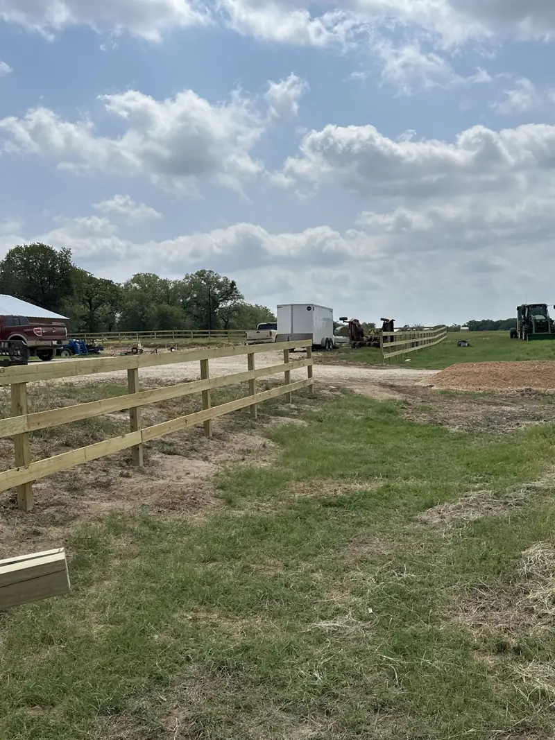 New three-board wood fence along a gravel drive, with a tractor and a second fence line in the distance.