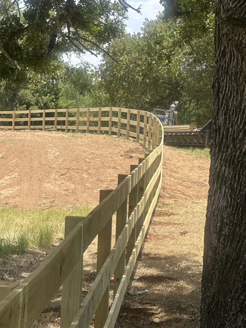 New wood board fence curving along a cleared dirt field, with a trailer of lumber parked behind it.