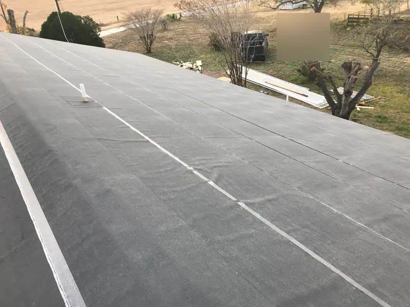 Roof covered in gray synthetic underlayment with taped seams, pasture beyond.