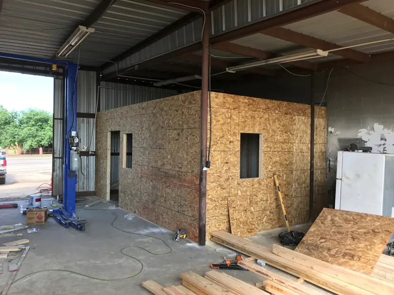 Office box sheathed in OSB inside a metal shop, next to a blue car lift.