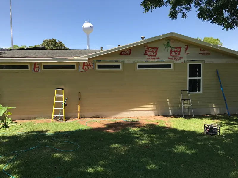 Long side of the house with a row of high windows and new lap siding, a water tower beyond the trees.
