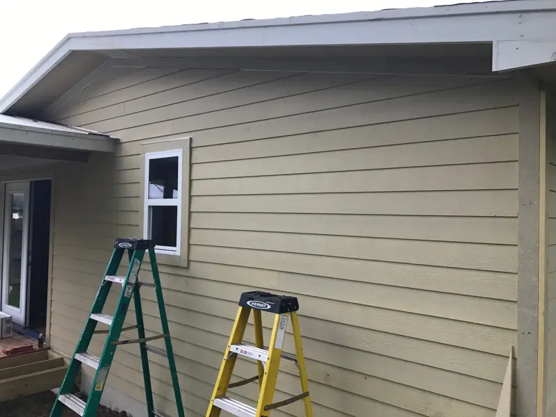 Corner of the house in new tan lap siding with a white window, ladders against the wall.