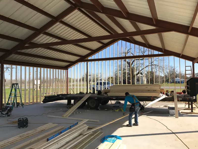 Wood wall studs going up inside an open steel building, with lumber on a trailer and cut boards on the slab.