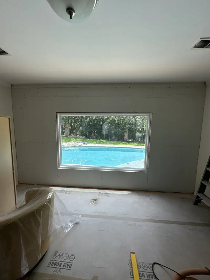 New window set in a freshly hung drywall wall, floor still covered in protection board.